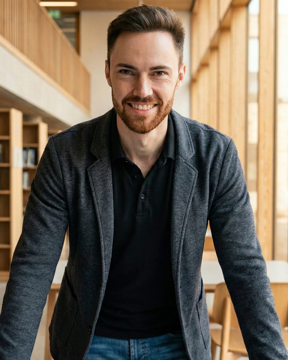 Robert Weller, smiling, leaning on a table in a bright library with wooden shelves